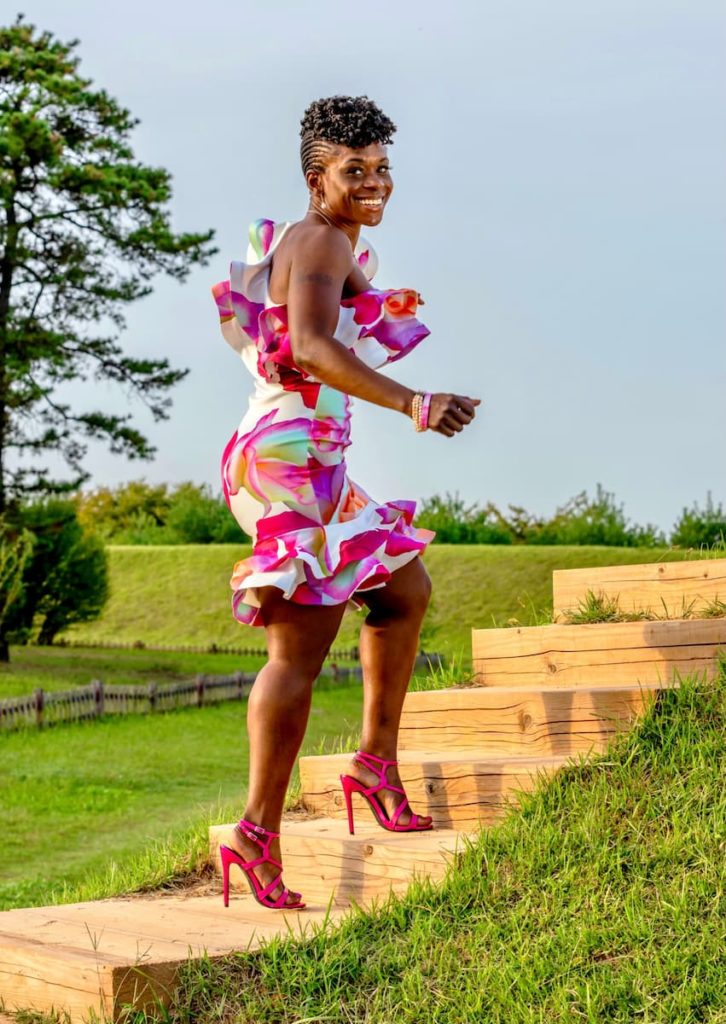 Octavia Scott, climbing some wooden stairs amidst grass, looking back over her shoulder. She is wearing a multicolor floral peplum dress and hot pink strappy shoes.
